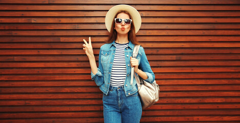 Portrait of young woman blowing her lips sending kiss wearing summer straw hat, backpack and jean jacket on wooden background