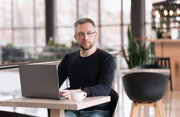 Fototapeta premium Successful adult man with glasses works on a laptop in a cafe