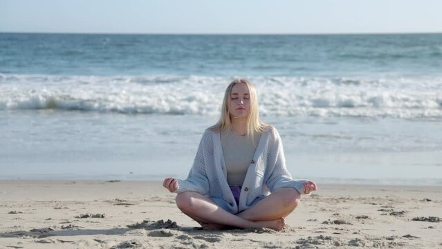 Blonde girl meditating on a sandy beach, washed with the calm ocean. Beautiful young woman sitting with closed eyes with the sunset sky in the background. High quality 4k footage