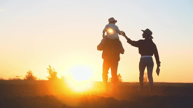 Silhouette of family:child is sitting on dad shoulders and mom walking next on summer evening at sunset in field under summer sun. Boy holds hands plucked wildflowers ,farmer holds digital tablet.