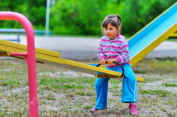 Young happy girl is swinging on the playground. Foliage of green plants is blurred in the background. A lot of midges fly around the child. Selective focus.