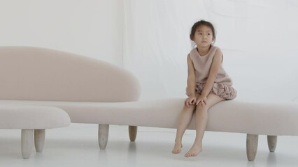 Cute Asian baby girl is sitting in a white room on the couch and dreaming, slow motion