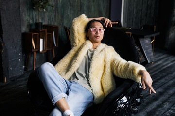 Portrait of Asian man in extravagant outfit with neon fur coat lounging in leather armchair at dark...