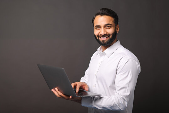 Indian male entrepreneur carrying laptop isolated on black, bearded businessman using laptop, typing, receiving emails, using new computer app, looking at the camera and smiles