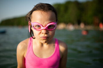Portrait of little girl in pink swimsuit and goggles making faces standing in water in lake in...