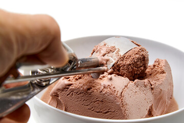 The hand of a person with an ice cream spoon scoops chocolate ice cream from a white bowl