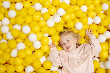 Fototapeta na wymiar Funny cute little caucaisian blonde baby girl,toddler, smiling kid having fun in ball pool,playing with yellow white balls.Happy child looking at camera in playground,top view.Copy space