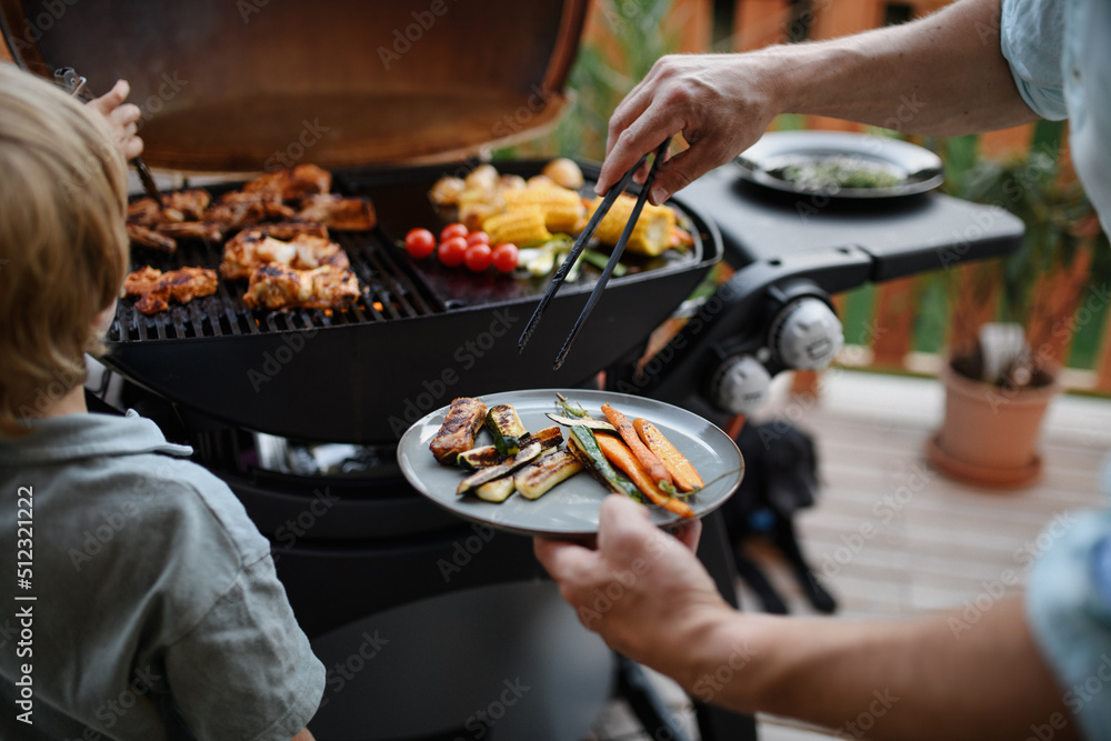 Wall mural unrecognizable father with little son grilling ribs and vegetable on grill during family summer gard