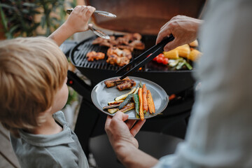 Unrecognizable father with little son grilling ribs and vegetable on grill during family summer...