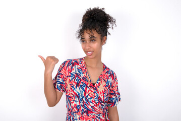 Stupefied Young African American woman wearing colourful dress with surprised expression, opens eyes and mouth widely, points aside with thumb, shows something strange. Advertisement concept.