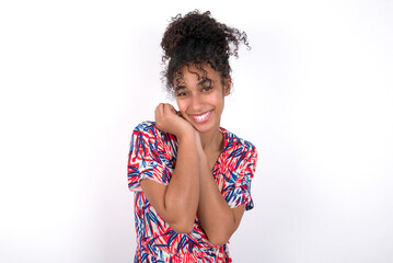 Dreamy Young African American woman wearing colourful dress over white wall with pleasant expression, closes eyes, keeps hands crossed near face, thinks about something pleasant