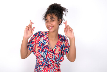 Joyful Young African American woman wearing colourful dress over white wall clenches teeth, raises fingers crossed, makes desirable wish, waits for good news, I have to win.