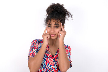 Young African American woman wearing colourful dress over white wall with thoughtful expression, looks away, keeps hand near face, thinks about something pleasant.