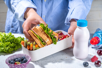 Back to school. Mother female hands making kids school lunch box with fresh fruits, berry, yogurt, healthy sandwich, vegetable snacks. White kitchen marble table copy