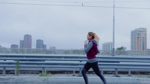 Young sportswoman running on sidewalk parallel to road. Girl is on morning jogging through streets of city. Cardio workout, athletics, sports activities. Woman, athlete training for marathon outdoor