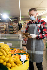 Portrait of staff using digital tablet while checking fruits in organic section of supermarket, young man grocery clerk in mask