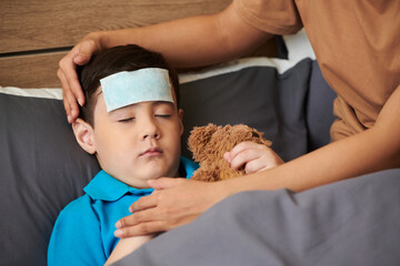 Sick child hugging teddy bear when sleeping in bed with cooling patch on forehead