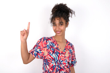 Young African American woman wearing colourful dress over white wall showing and pointing up with fingers number one while smiling confident and happy.