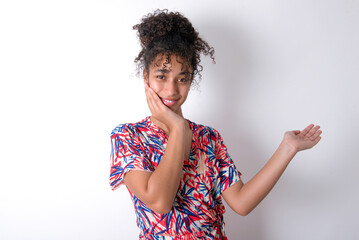 Positive glad Young African American woman wearing colourful dress over white wall says: wow how exciting it is, indicates something.  One hand on his head and pointing with other hand.