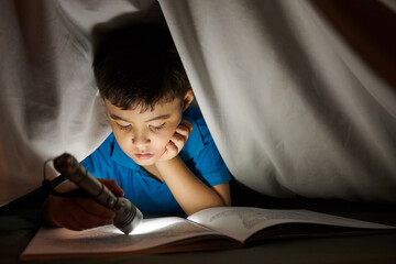 Preteen boy using flashlight when reading book under blanket late at night