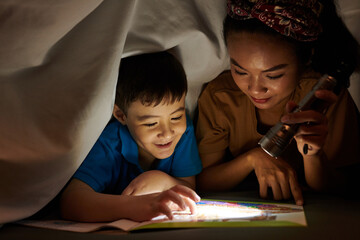 Mother shining flashlight on book page when son reading scary story - obrazy, fototapety, plakaty