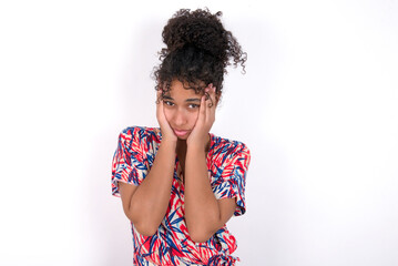 Young African American woman wearing colourful dress over white wall holding head in hands with unhappy expression watching sad movie about animals and trying not to cry.