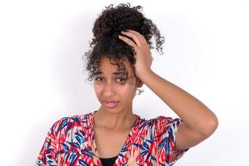 Portrait of confused Young African American woman wearing colourful dress over white wall holding...