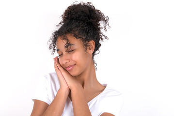 Fototapeta na wymiar Young beautiful girl with afro hairstyle wearing white t-shirt over white wall leans on pressed palms closes eyes and has pleasant smile dreams about something