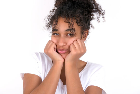 Young beautiful girl with afro hairstyle wearing white t-shirt over white wall with surprised expression keeps hands under chin keeps lips folded makes funny grimace