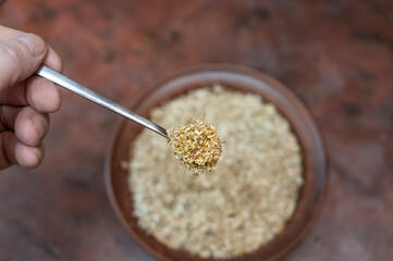 Grown man's hand holds full spoonful of dried Matricaria chamomilla flowers over the kitchen table. A brown bowl of medicinal herbs blurred in the background. Selective focus. Series part.