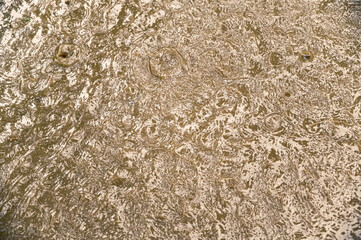 Texture violent rain stream on the city street. A torrential downpour on paved road. Muddy yellow water flows down to storm drains. Falling large raindrops with splashes. Selective focus.