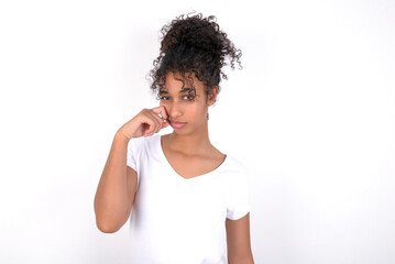 Disappointed dejected Young beautiful girl with afro hairstyle wearing white t-shirt over white wall wipes tears stands stressed with gloomy expression. Negative emotion