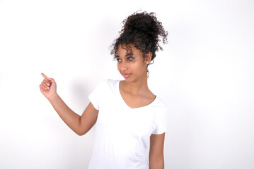 Young beautiful girl with afro hairstyle wearing white t-shirt over white wall points at copy space and advertises something, advices best price.