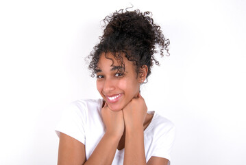 Young beautiful girl with afro hairstyle wearing white t-shirt over white wall holds hands under chin, glad to hear heartwarming words from stranger