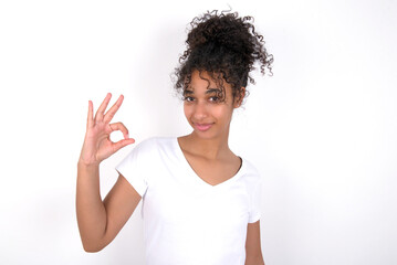 Young beautiful girl with afro hairstyle wearing white t-shirt over white wall hold hand arm okey symbol toothy approve advising novelty news