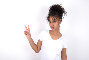 Young beautiful girl with afro hairstyle wearing white t-shirt over white background makes peace gesture keeps lips folded shows v sign. Body language concept