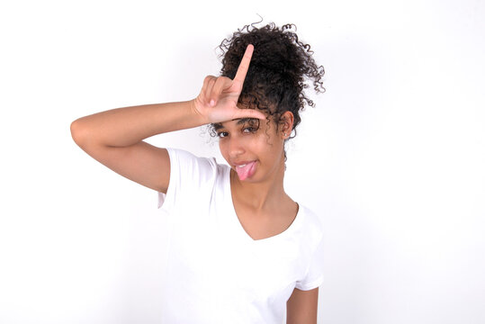 Funny Young beautiful girl with afro hairstyle wearing white t-shirt over white background makes loser gesture mocking at someone sticks out tongue making grimace face.