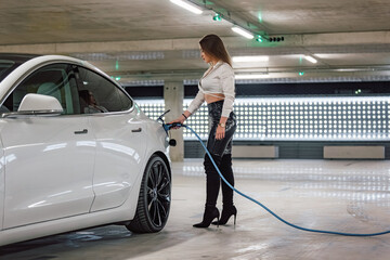 Attractive young woman charging a luxury electric car at an electric vehicle charging station in a...