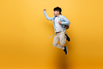Fototapeta na wymiar Side view of asian schoolkid with backpack jumping on yellow background.