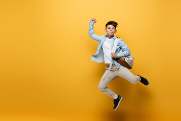 Fototapeta na wymiar Asian schoolkid holding backpack and showing yes gesture while jumping on yellow background.