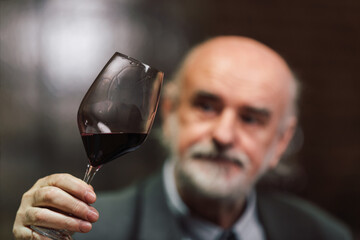 Caucasian senior man in a suit, standing in an old wine cellar with wooden barrels, expertly tasting red wine, checking a color, smell, and taste