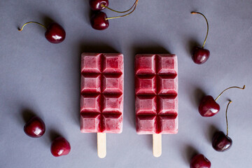 Naklejka na ściany i meble Homemade berry ice cream or popsicle sorbet on a stick. Two pieces on a gray background, cherries are laid out around.