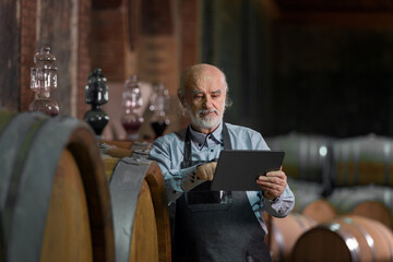 Caucasian senior man, a graybeard winemaker with apron checking big wooden barrels in a wine cellar and entering data in a tablet device