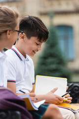 Smiling asian schoolboy holding pen and notebook near blurred friend outdoors.