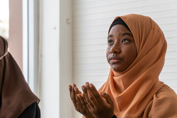 A young black Muslim girl in a brown hijab praying looking up and raise her hands A calm face in a white room
