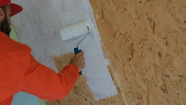 a worker with a beard in an orange robe is painting osb plate on the ceiling in white color