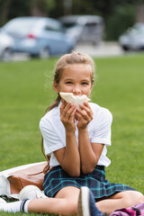 Schoolgirl holding sandwich near face on grass in park.