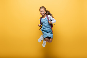 Happy preteen schoolgirl with backpack jumping on yellow background.