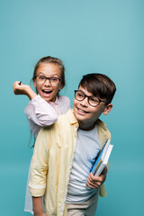 Cheerful schoolgirl in eyeglasses holding pen near asian classmate with notebooks isolated on blue.