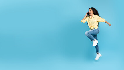 Fototapeta na wymiar Excited young woman talking on mobile phone, making call, jumping and running on yellow background, blank space